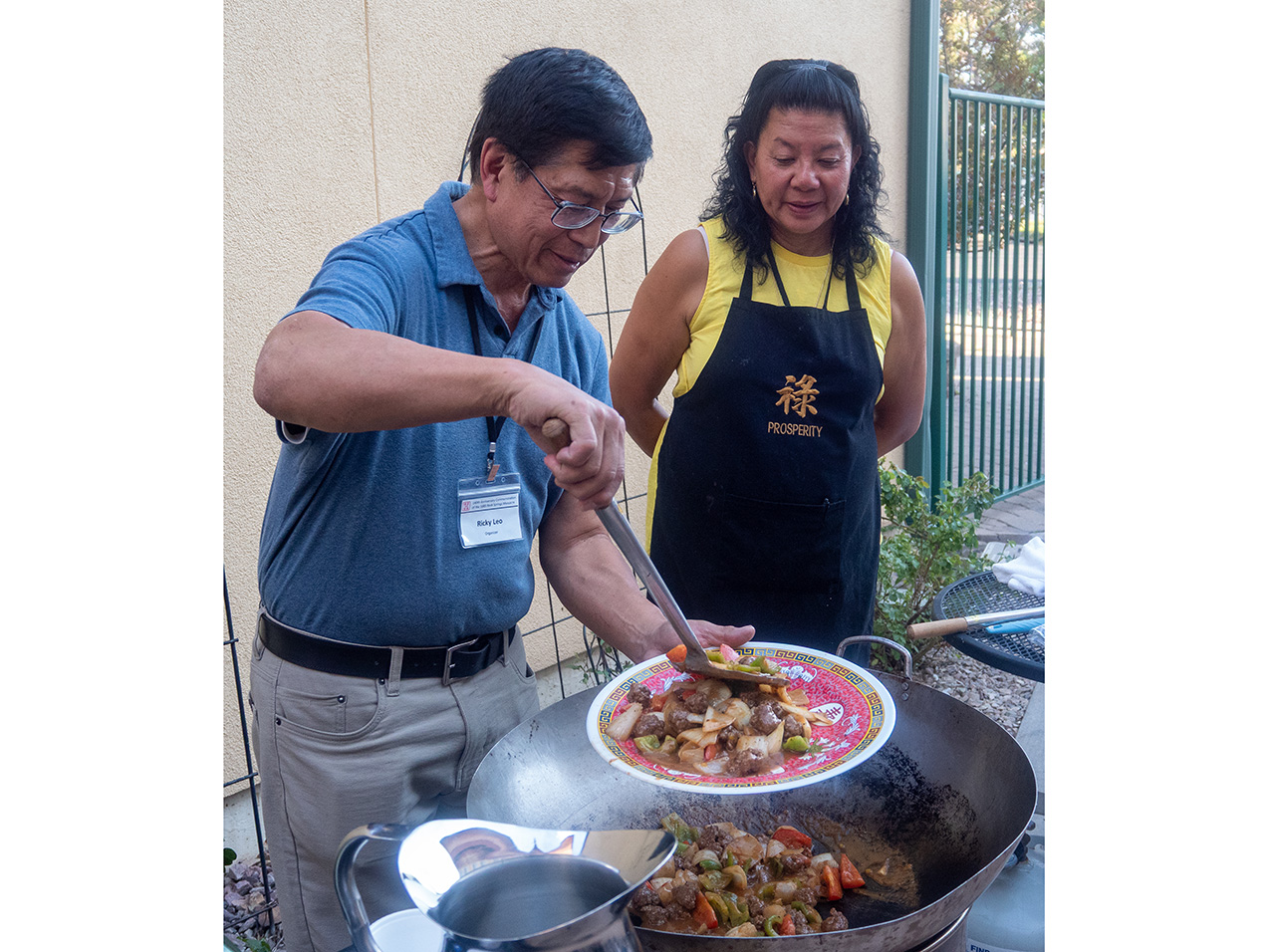 in front of a stucco wall and fence, an Asian man in khakis and a blue shirt, with glasses and a nametag that says RIcky Leo, uses a spatula to scoop vegetables and meat from a large wok onto a Chinese patterned plate. an Asian woman stands next to him, wearing a yellow top and a black apron.