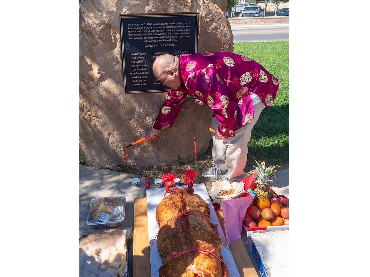 a bald-headed Chinese man, wearing a magenta silk shirt with an Asian pattern, bends down to light candles that have been set in front of a rock memorial. In the foreground are offerings of food such as sliced chicken, a pineapple and other fruits, and a large roasted animal tied to a board.
