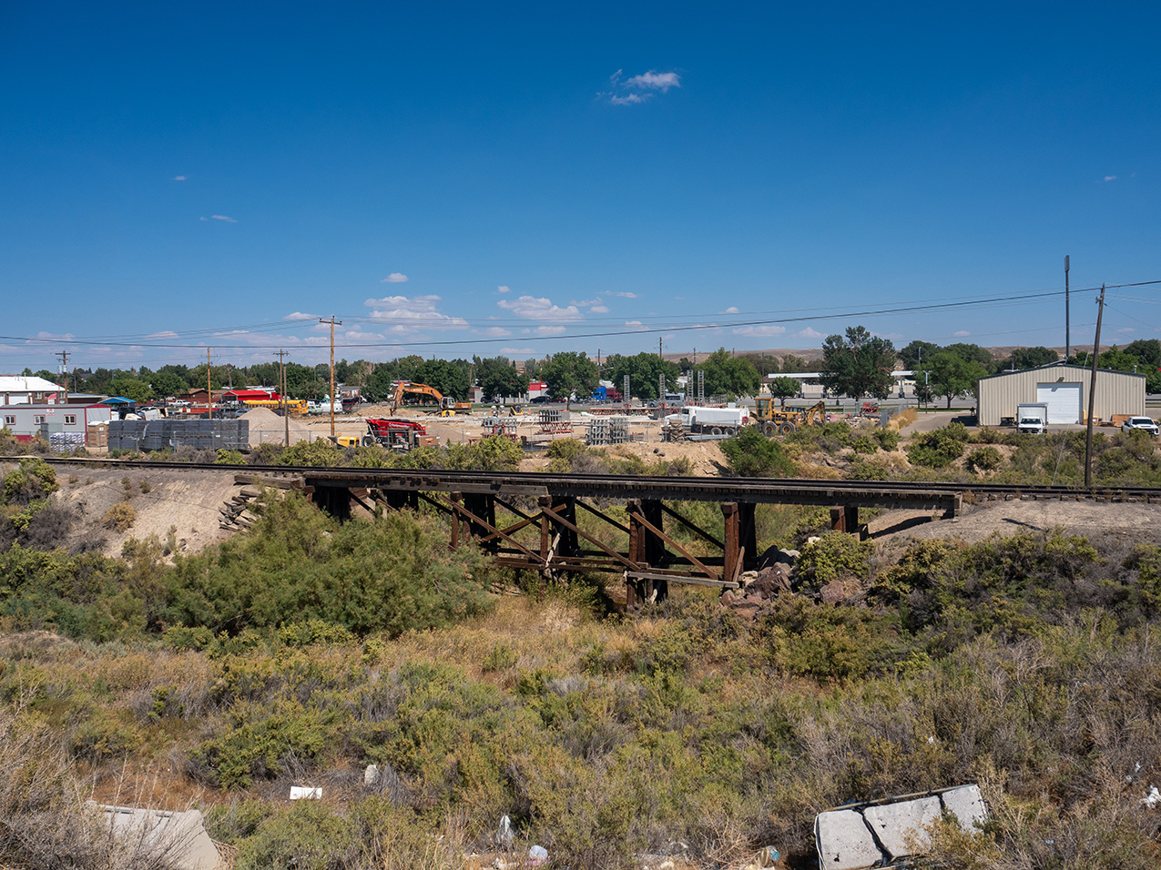 a railroad trestle spans a low-lying scrubby area with some litter among the plants. the background is an industrial area with buildings and heavy equipment.