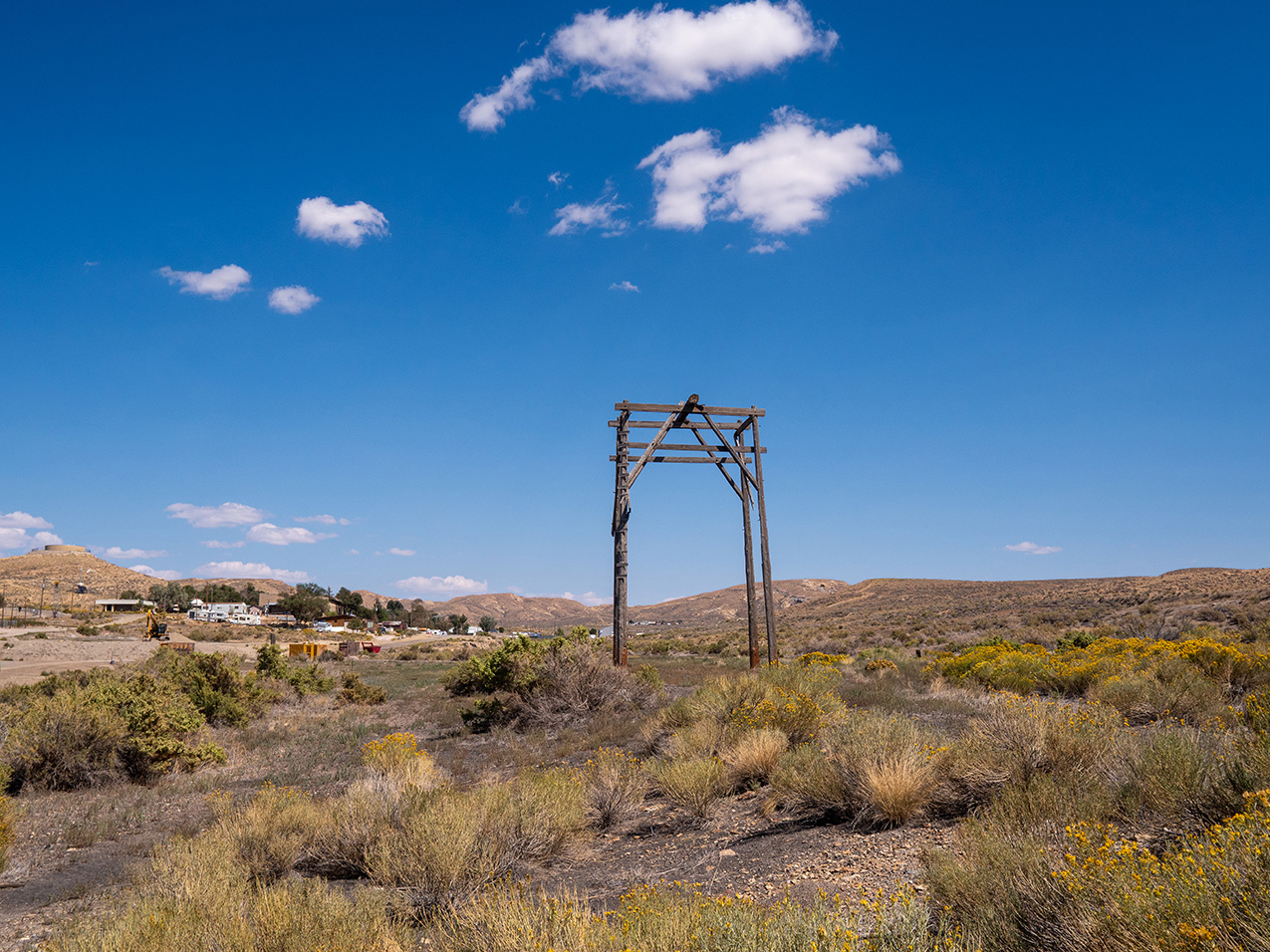 a tall wooden structure stands alone in a field of short scrubby plants. the background features low rolling hills and hints of human settlement.