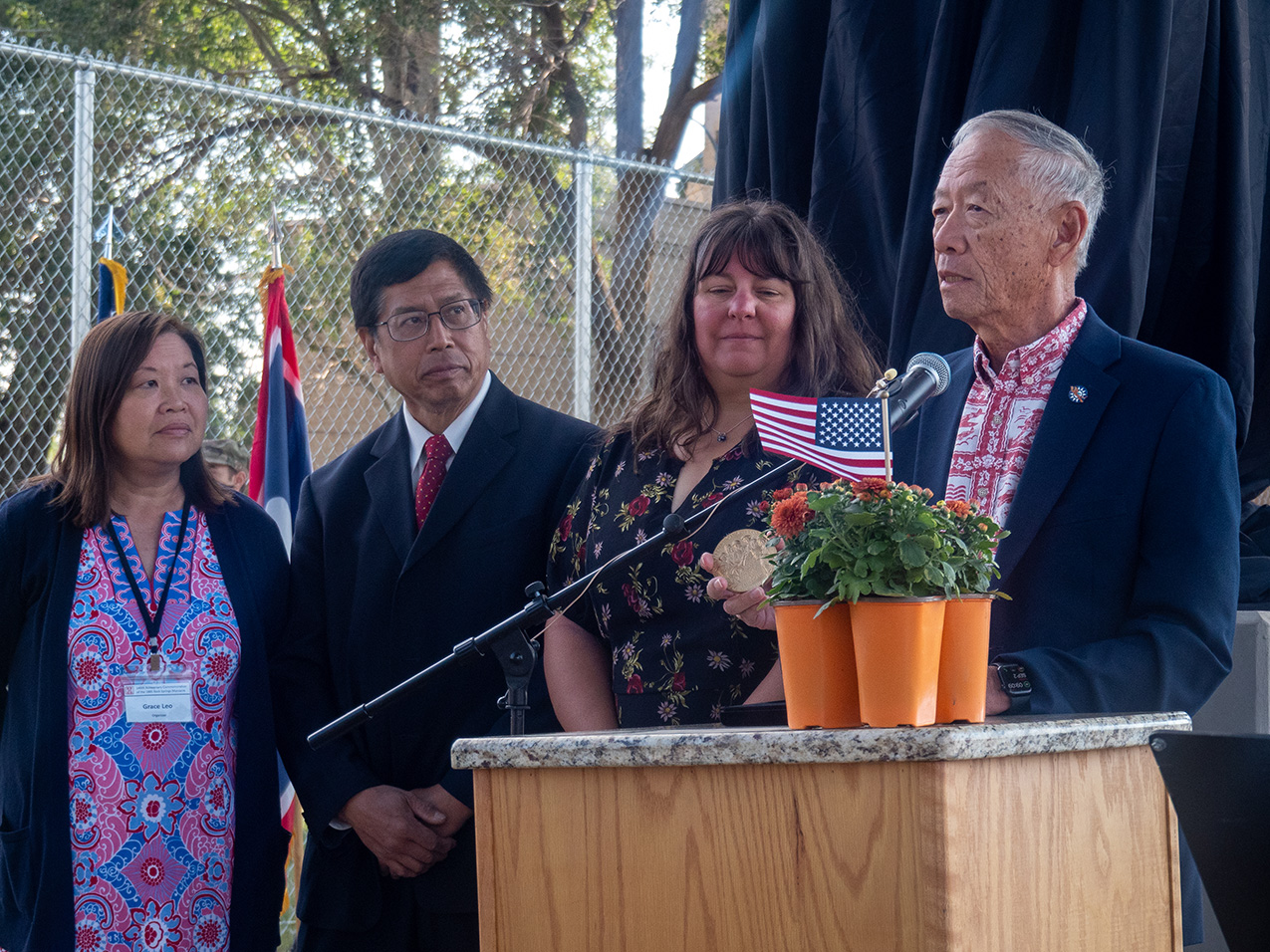 a Chinese man, holding a round medal, speaks into a microphone at a podium with a small American flag and orange flowered plants in bright orange pots. three other people, standing in close proximity to him, look at his face or the medal.