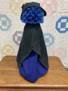 a textile item resembling a primitive doll: a pompom of blue yarn for the head, black fabric for a hat, a black washcloth for a shawl, and blue fabric beneath the shawl. It sits on a wooden pedestal in front of a hanging quilt backdrop.